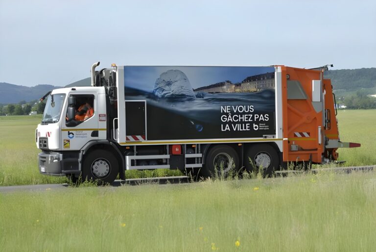 A Grand Besançon Métropole waste collection truck with a Sulo body driving past a grassy field.