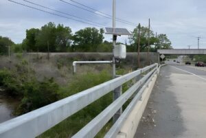 Solar-powered Green Stream Technologies flood sensor mounted on a bridge guardrail overlooking a creek.