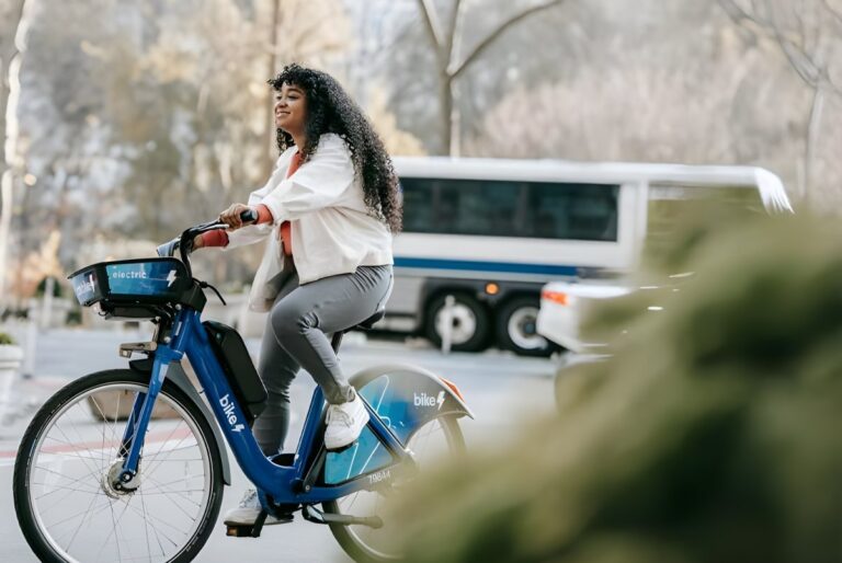 A woman rides a blue Bay Wheels electric bike on a city street with a white transit bus blurred in the background.