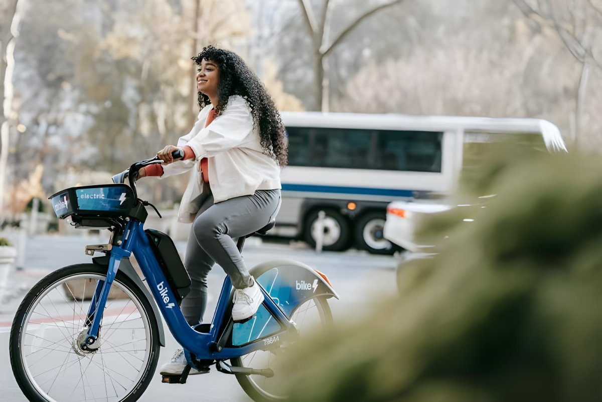 A woman rides a blue Bay Wheels electric bike on a city street with a white transit bus blurred in the background.