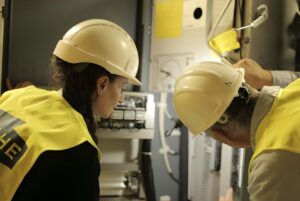 Two HEDNO technicians in hard hats and yellow safety vests inspect an electrical panel and utility meters.