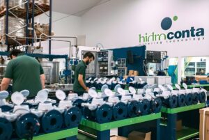 Two workers assemble rows of blue smart water meters in a Hidroconta factory with branded wall signage.