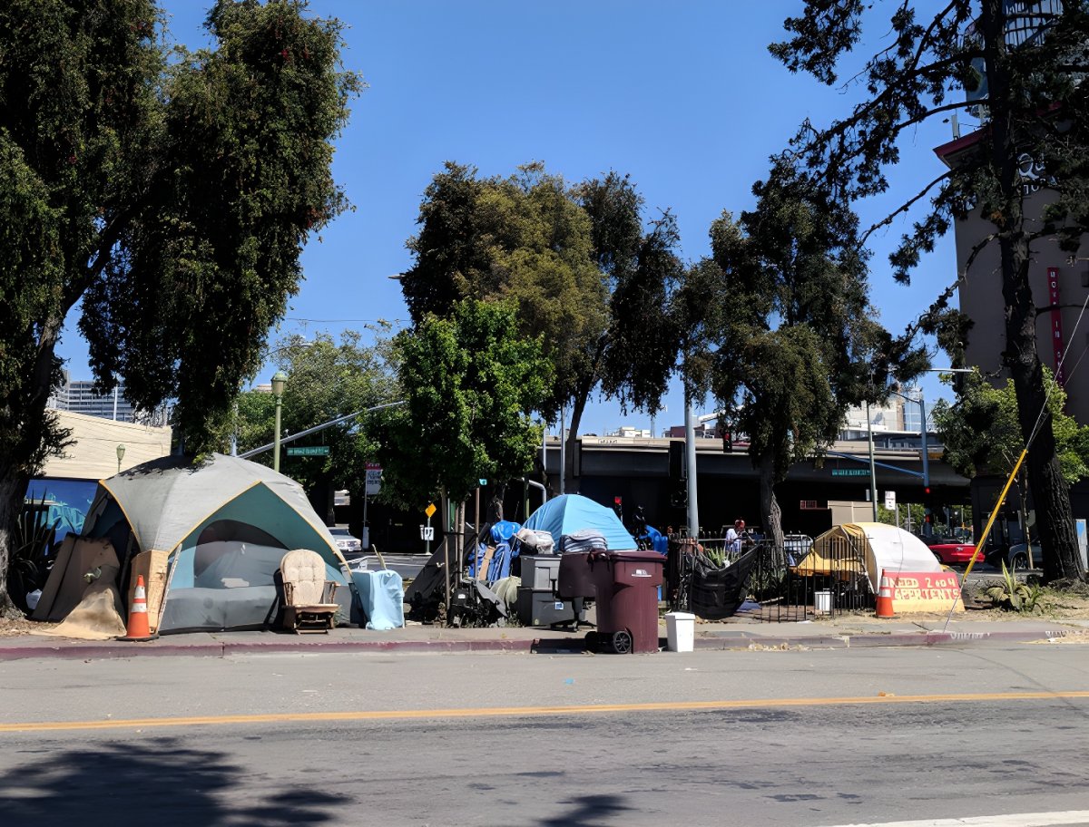 Tents, trash bins, and personal belongings in a homeless encampment on a city sidewalk under trees near an overpass.