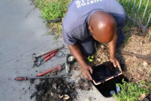 A Houston Public Works employee uses a tablet to inspect an underground water meter box surrounded by hand tools.