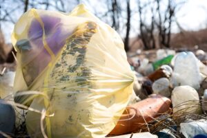 Close-up of a yellow plastic bag filled with waste sitting on a pile of discarded bottles and litter in a wooded area.