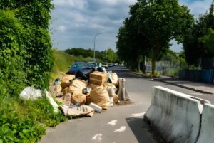 Large pile of illegally dumped trash bags and cardboard boxes on a paved roadside next to a concrete barrier.