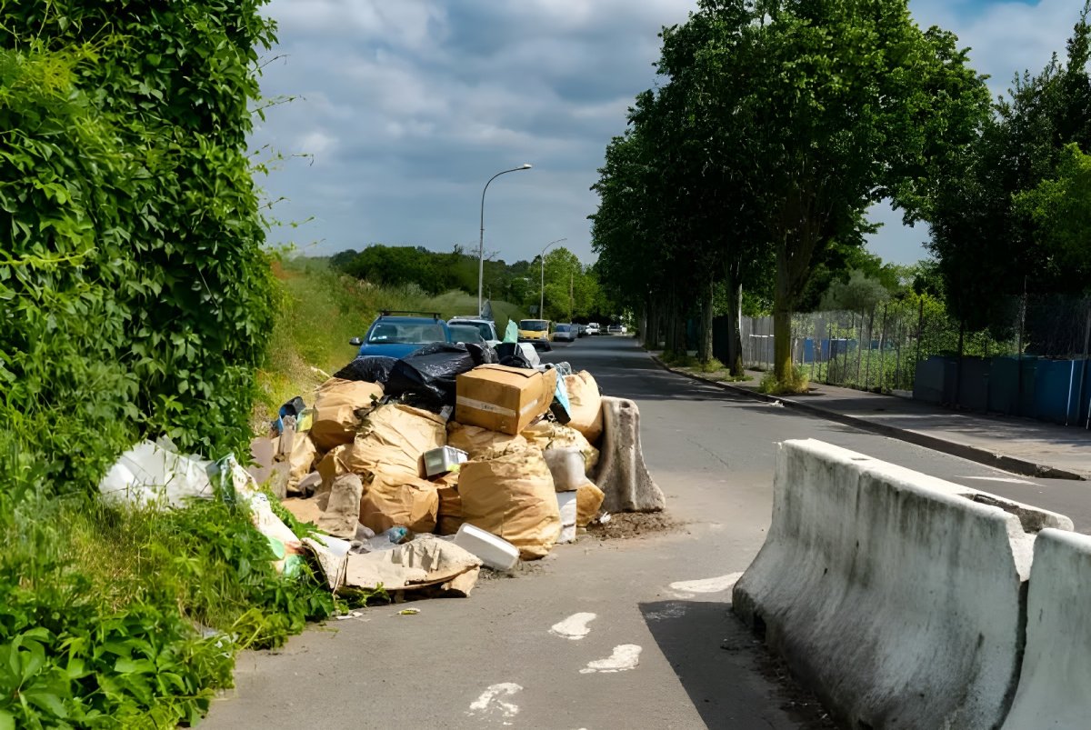 Large pile of illegally dumped trash bags and cardboard boxes on a paved roadside next to a concrete barrier.