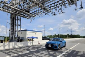 Blue Audi driving under an Indra overhead tolling gantry with sensors and cameras on a highway.