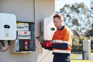 Technician in high-visibility clothing using a tablet while inspecting an outdoor electrical meter box and solar inverters.