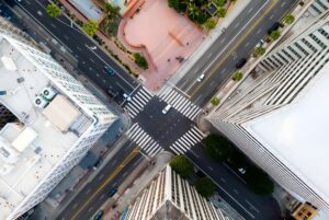 Top-down aerial view of a four-way city intersection with crosswalks, moving vehicles, and surrounding office buildings.