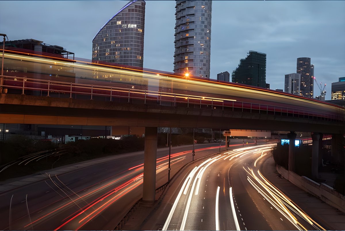 Long exposure of light trails from vehicles on a multi-lane highway and elevated train track in a city at dusk.