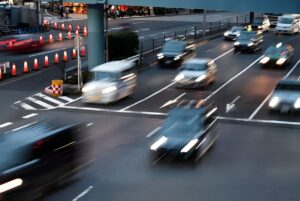Motion-blurred cars with headlights on driving through a multi-lane city intersection at dusk.