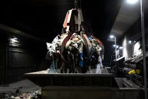 Industrial orange peel grabber lifting a load of mixed waste inside a dark waste-to-energy facility.
