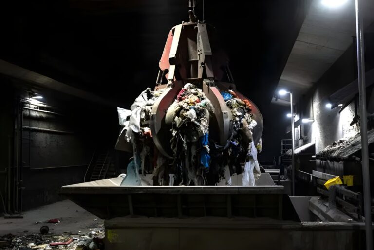 Industrial orange peel grabber lifting a load of mixed waste inside a dark waste-to-energy facility.