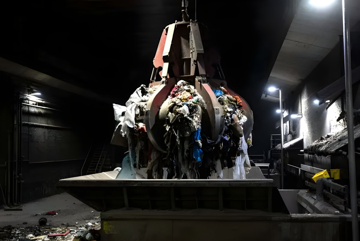 Industrial orange peel grabber lifting a load of mixed waste inside a dark waste-to-energy facility.