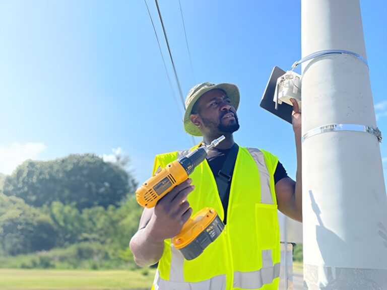 Technician in a high-visibility vest using a power drill to install a JustAir monitoring device on a concrete utility pole.