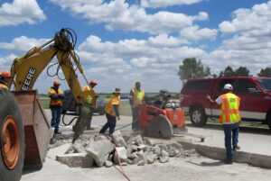 Construction crew using a John Deere excavator and concrete saw to repair a road under a cloudy sky.