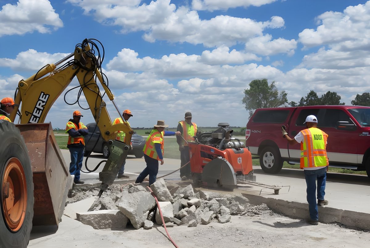 Construction crew using a John Deere excavator and concrete saw to repair a road under a cloudy sky.