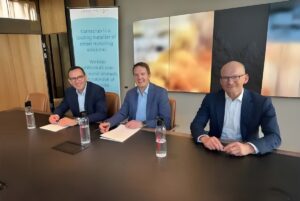 Three men in suits sit at a conference table with documents and water bottles in front of a Kamstrup branded banner.