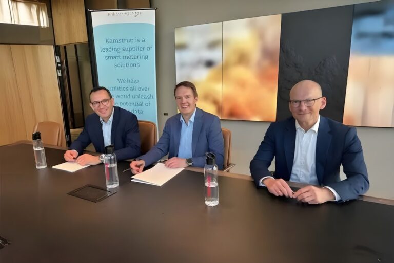 Three men in suits sit at a conference table with documents and water bottles in front of a Kamstrup branded banner.