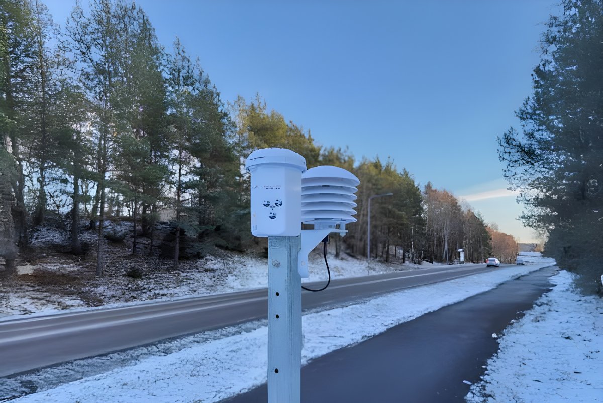 Klimator IoT road weather station mounted on a pole beside a snow-dusted highway lined with evergreen trees.