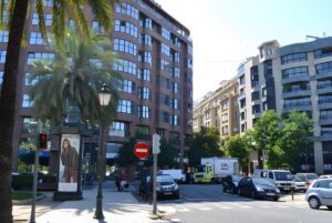 Street-level view of a city intersection with multi-story apartment buildings, palm trees, and traditional street lamps.