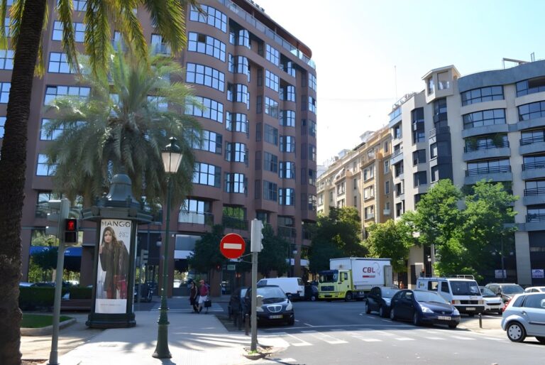 Street-level view of a city intersection with multi-story apartment buildings, palm trees, and traditional street lamps.