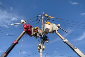 Utility workers in two bucket truck lifts repair electrical components on a power pole against a blue sky.