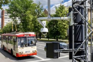 White dome security camera mounted on a metal pole overlooking a red and cream trolleybus on a city street.