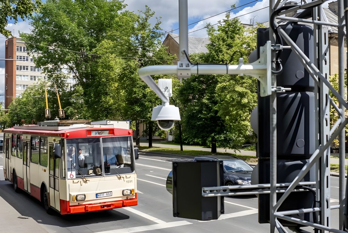 White dome security camera mounted on a metal pole overlooking a red and cream trolleybus on a city street.