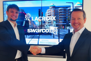 Two men in suits shake hands in front of a screen displaying the LACROIX and SWARCO logos over a city street scene.