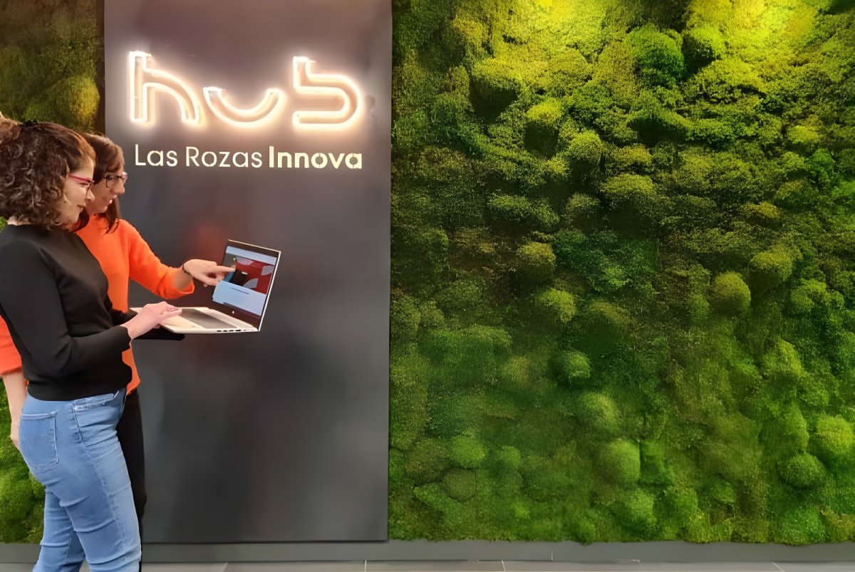 Two women use a laptop in front of a Las Rozas Innova hub sign and a green moss wall.