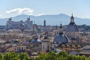 Panoramic view of the Rome skyline featuring the Altare della Patria and various church domes against distant mountains.