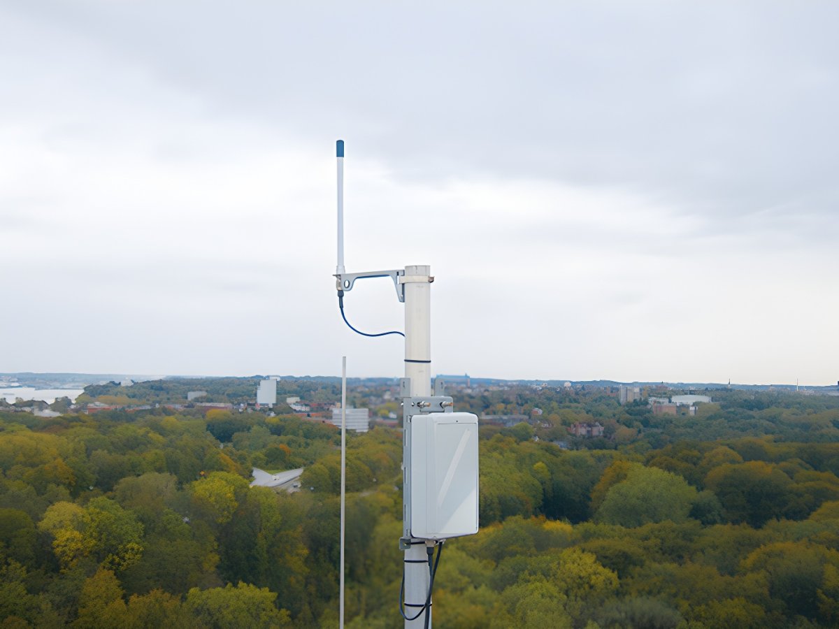 LoRaWAN gateway and antenna mounted on a metal pole overlooking a forested landscape and distant city buildings.