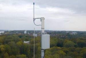 LoRaWAN gateway and antenna mounted on a rooftop pole overlooking a forested landscape and distant town.