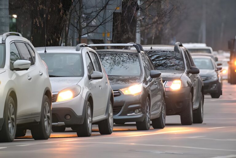 Line of cars with headlights on driving in heavy city traffic during the day.