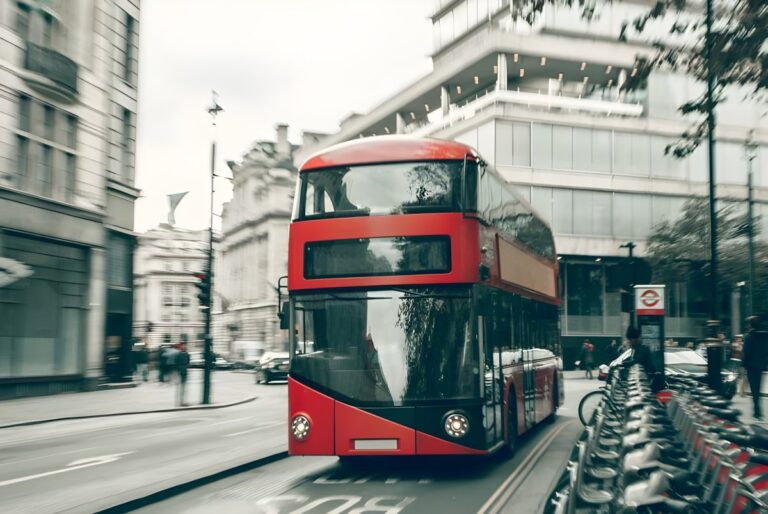 Red double-decker bus driving on a London street past a row of docked public bicycles and multi-story buildings.