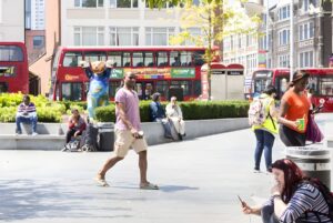 Pedestrians walking through a London plaza with red double-decker buses and a Paddington Bear statue in the background.