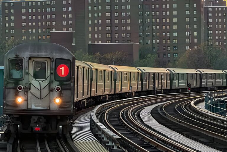 An MTA New York 1 train travels along an elevated curved track past brick apartment buildings.