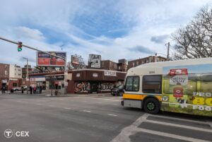 An MBTA bus drives through a Boston city intersection with traffic lights, billboards, and storefronts under a blue sky.
