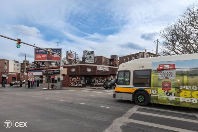 An MBTA bus drives through a Boston city intersection with traffic lights, billboards, and storefronts under a blue sky.