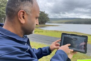 A man in a blue jacket uses a tablet to monitor a reservoir surrounded by forest.