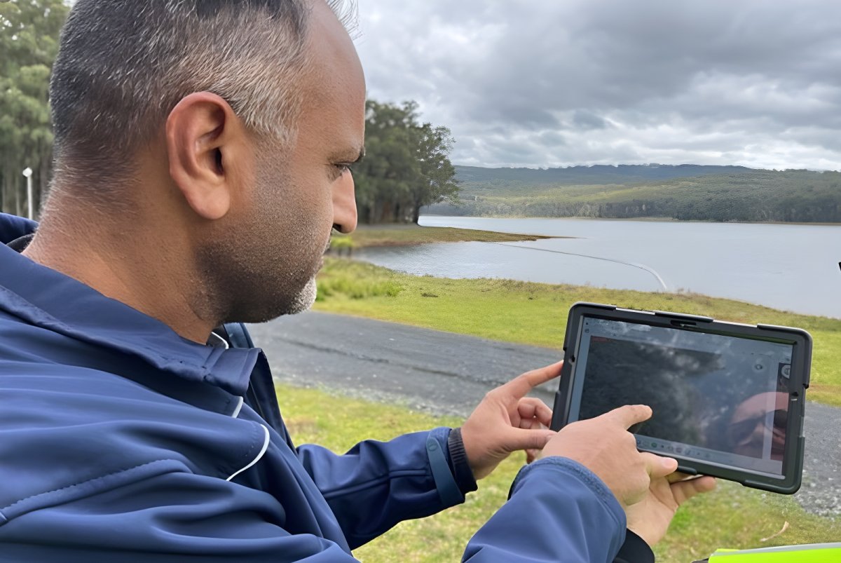 A man in a blue jacket uses a tablet to monitor a reservoir surrounded by forest.