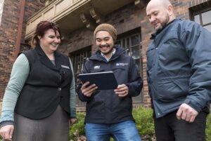 Three people stand outside a brick building while a man in a Melbourne Water jacket holds a digital tablet.