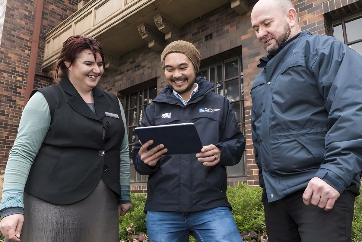 Three people stand outside a brick building while a man in a Melbourne Water jacket holds a digital tablet.