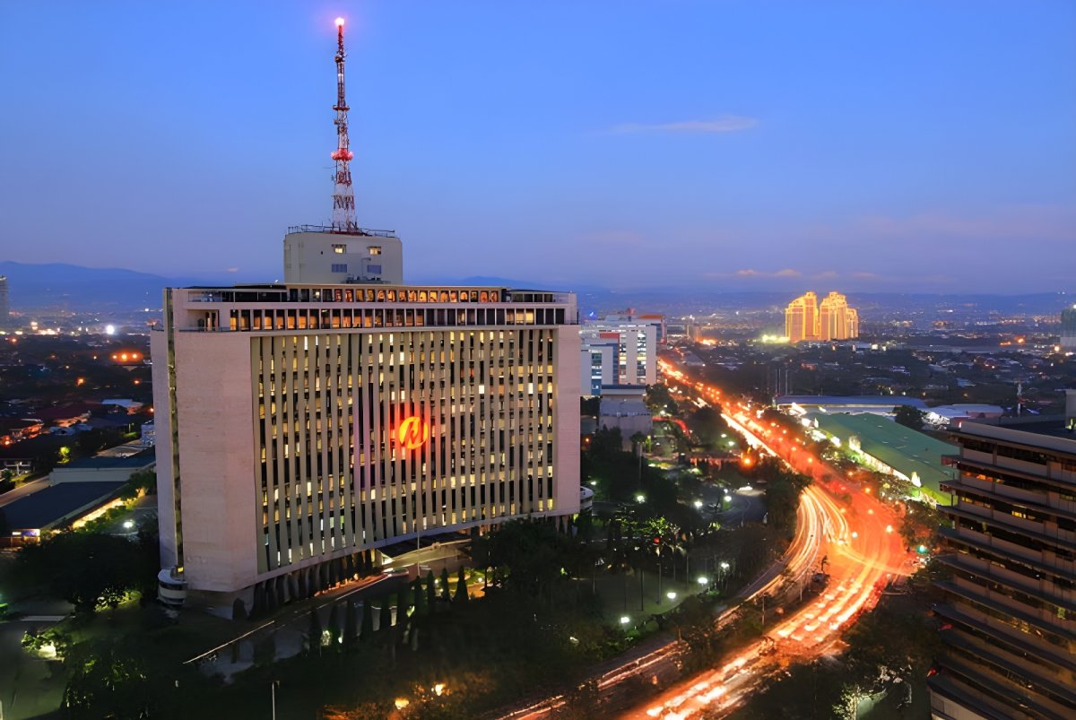 The Meralco headquarters building at dusk with a lit logo, telecommunications tower, and city traffic light trails.