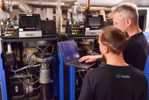 Two technicians in Metr branded shirts use a laptop to configure industrial Buderus heating boilers in a utility room.