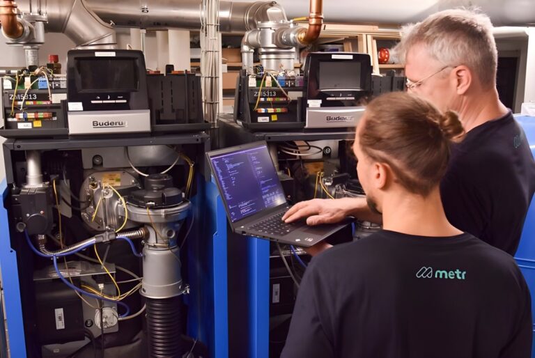 Two technicians in Metr branded shirts use a laptop to configure industrial Buderus heating boilers in a utility room.