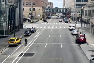 High-angle view of a city street with a dedicated bike lane, parked cars, and a pedestrian skyway bridge in the distance.