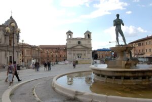 Pedestrians walk through Piazza Duomo in L'Aquila past the Fontana Vecchia and street lighting poles.
